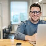 Happy man wearing glasses working remotely on laptop in modern office environment.